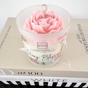 May include: A pink peony-shaped candle in a clear glass jar. The jar has floral designs and the text "Peony Blossom" and "Hand Poured Cocosoy Floral Candle". The candle sits on a book with the text "at 100 CENTURY OF STYLE".