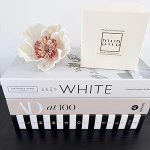 May include: A cream-coloured decorative flower with a reddish-brown centre sits on a stack of books. A white box labelled "Premium Diffuser Flowers" is also present. The books are titled "WHITE", "AD at 100", and "ELEMENTS OF STYLE".