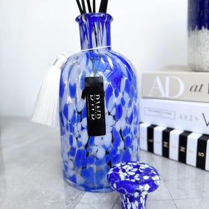 May include: A blue and white speckled glass diffuser bottle with black reeds and a white tassel. A matching mushroom-shaped stopper sits in front. The bottle has a black label with white text. Decorative home fragrance item.