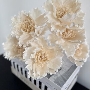 May include: A collection of cream-coloured artificial peony flowers with ruffled petals and textured centres. The flowers sit on a stack of books with black and white striped spines and the text "CHRISTIANA NORA".