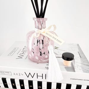 May include: A pink and white speckled glass diffuser bottle with black reeds, a white tassel, and a small jar with a black lid. The bottle is tied with a white ribbon and a tag that reads "BELLA'S WAX BARN". The items are arranged on top of books.