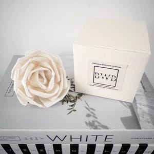 May include: A cream-coloured paper rose and a white box labelled "PREMIUM DIFFUSER FLOWERS" with the brand "BWD" sit on books. The books have the word "WHITE" on the spine. The box and rose are on a light grey surface.