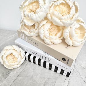May include: Ivory peony flowers rest on a stack of books. The books are in neutral tones, with the top book having the text "AD at 100" and the middle book displaying the word "WHITE". The overall aesthetic is soft and elegant.