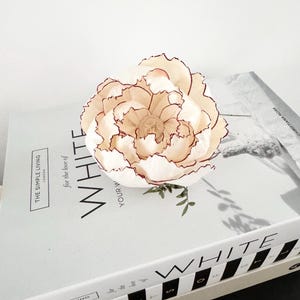 May include: A cream-coloured artificial flower with dark red edges rests on a stack of white books. The flower has multiple layers of petals and a detailed centre. The books have the words "WHITE" and "THE SIMPLE LIVING" printed on them.