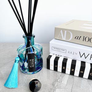 May include: A decorative glass diffuser bottle with a blue and teal swirl design, black reeds, and a turquoise tassel. A black cap sits next to the bottle. Books with the text "AD at 100", "THE SIMPLE LIVING", and "ELEMENTS" are stacked in the background.