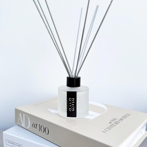 May include: A frosted glass diffuser bottle with a black cap and grey reeds. The bottle has a black and white label that reads "DIAUD". The diffuser sits atop a stack of books, including one titled "AD at 100".