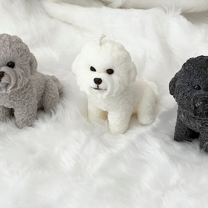 May include: Three small dog-shaped candles, one grey, one white, and one black, are displayed on a white fluffy surface.