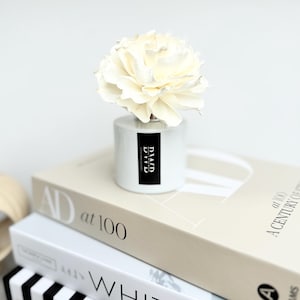 May include: A white ceramic essential oil diffuser with a large, cream-coloured flower on top. The diffuser has a black and white label with the text "DVD". The diffuser sits on top of a stack of books.
