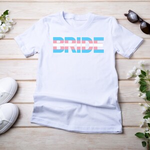 Könnte beinhalten: Weißes T-Shirt mit dem Wort "BRIDE" in blauen Buchstaben mit einem Transgender-Pride-Flaggen-Design.