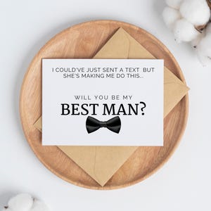 May include: A white card with black text that reads "I could've just sent a text but she's making me do this... Will you be my Best Man?" with a black bow tie illustration.