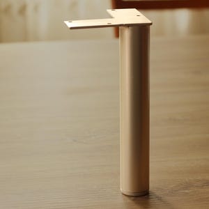 Könnte beinhalten: Ein zylindrisches Möbelbein mit einer metallischen champagnerfarbenen Oberfläche. Das Bein verfügt über eine flache, L-förmige Montageplatte an der Oberseite mit vier vorgebohrten Schraublöchern zur Befestigung an einer ebenen Fläche.
