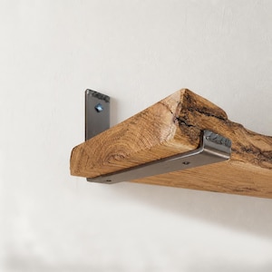 Peut inclure: Une étagère murale composée d'une épaisse planche en bois brut avec un bord naturel et un grain apparent. L'étagère est fixée par un support métallique en forme de L, de couleur sombre, qui se fixe au mur et soutient le dessous du bois.
