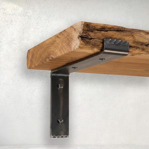 Op de afbeelding: Een houten plank met een natuurlijke boomkant, zichtbare houtnerf en een ruwe voorzijde. De plank wordt ondersteund door een donkere, robuuste L-vormige metalen beugel met een gehamerde afwerking, vastgezet met zichtbare bouten.