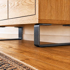 May include: A close-up of a wooden cabinet with a light brown finish, supported by dark gray, rectangular metal legs. The legs are attached to the bottom of the cabinet. The cabinet sits on a wooden floor.