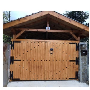 May include: A double-door wooden gate featuring vertical light brown planks, black metal hinges, and decorative black studs. A black metal ring-shaped door knocker is centered on the right panel, and a small square sign is mounted above.