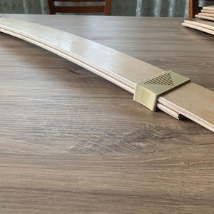 May include: A light-coloured wooden leaf spring with a brass-coloured metal bracket. The spring is curved and appears to be made of multiple layers of wood. The bracket has a geometric design.