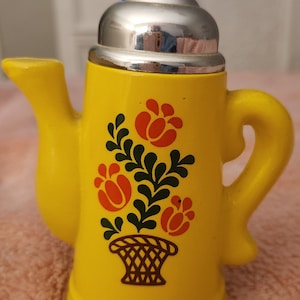 Puede incluir: Una tetera de cerámica amarilla con tapa y asa plateadas. La tetera está decorada con un diseño floral en rojo, verde y marrón. Tiene un pico y un asa. Es probable que sea un objeto decorativo.