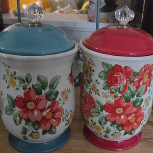 May include: Two ceramic canisters with cream-colored bodies featuring floral patterns of red and yellow flowers with green leaves. One canister has a blue lid and base, while the other has a red lid and base. Both feature clear glass knobs on top.