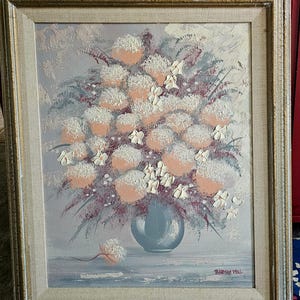 May include: Oil painting of a floral arrangement in a blue vase, set against a light grey background. The flowers are a mix of peach and white, with green and purple accents. The painting is in a gold-toned frame, signed "THOMAS PAL".