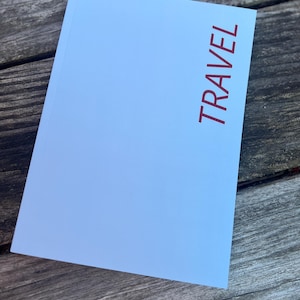 May include: A light blue notebook with the word "TRAVEL" printed vertically in red. The notebook is lying on a weathered wooden surface.