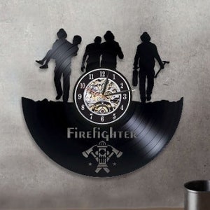 Puede incluir: Reloj de vinilo negro con temática de bomberos. El reloj presenta una silueta de cuatro bomberos y la palabra "Bombero" en letras blancas. La esfera del reloj es un reloj analógico tradicional con números romanos.