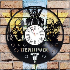Puede incluir: Un reloj de vinilo negro con una silueta de Deadpool en tres poses diferentes. La esfera del reloj es blanca con números romanos negros y una aguja de hora roja y una aguja de minutos negra. El reloj está rodeado de un fondo negro con la palabra "DEADPOOL" en letras blancas y un par de pistolas cruzadas.