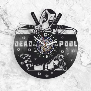 Puede incluir: Reloj de vinilo negro con una silueta de Deadpool en una pose de superhéroe. La esfera del reloj está en el centro del disco con las palabras "DEADPOOL" impresas en la parte superior e inferior de la esfera.