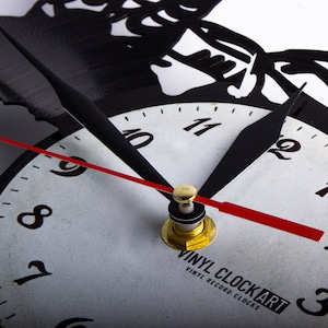 May include: A white clock face with black numbers and hands. The clock is made from a black vinyl record with a cut-out design. The clock face reads "VINYL CLOCKART VINYL RECORD CLOCKS".