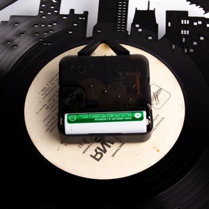 May include: A black vinyl record clock with a white face and black hands. The clock is powered by a single AA battery. The clock face has the text "QUARTZ" and "PLASTINOOK" printed on it.