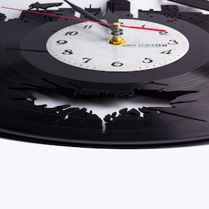 May include: A black vinyl record clock with a cityscape design. The clock face is white with black numbers and hands. The clock is broken in the shape of a bat.