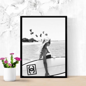 K&ouml;nnte beinhalten: Schwarzwei&szlig;foto einer Frau, die ein Surfbrett mit Chanel-Logo h&auml;lt. Die Frau tr&auml;gt einen Badeanzug und eine Sonnenbrille. Der Hintergrund ist ein Strand mit Palmen.