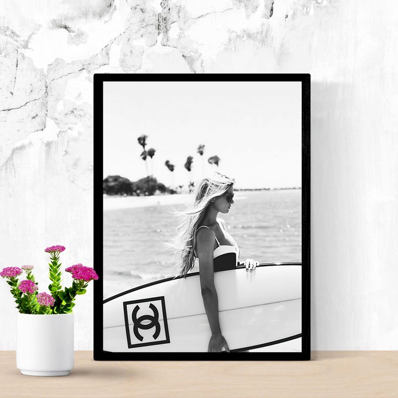 K&ouml;nnte beinhalten: Schwarzwei&szlig;foto einer Frau, die ein Surfbrett mit Chanel-Logo h&auml;lt. Die Frau tr&auml;gt einen Badeanzug und eine Sonnenbrille. Der Hintergrund ist ein Strand mit Palmen.