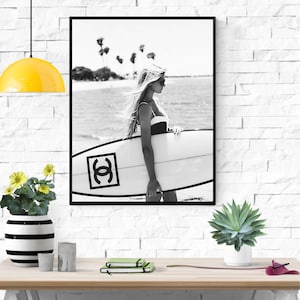 K&ouml;nnte beinhalten: Schwarzwei&szlig;foto einer Frau, die ein Surfbrett mit einem Chanel-Logo h&auml;lt. Die Frau tr&auml;gt einen Badeanzug und eine Sonnenbrille. Das Foto wurde an einem Strand mit Palmen im Hintergrund aufgenommen.