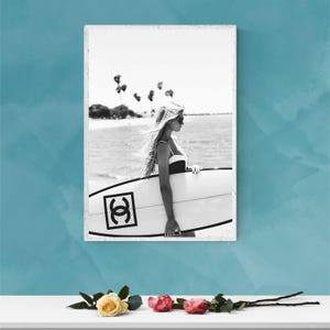 K&ouml;nnte beinhalten: Schwarzwei&szlig;foto einer Frau, die ein Surfbrett mit Chanel-Logo h&auml;lt. Die Frau tr&auml;gt einen Badeanzug und eine Sonnenbrille. Das Foto wurde an einem Strand mit Palmen im Hintergrund aufgenommen.