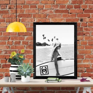 K&ouml;nnte beinhalten: Schwarzwei&szlig;foto einer Frau, die ein Surfbrett mit einem schwarzen "Chanel"-Logo an der Seite h&auml;lt. Die Frau tr&auml;gt einen Badeanzug und hat langes Haar. Das Foto ist in einem schwarzen Rahmen gerahmt und h&auml;ngt an einer Backsteinwand.