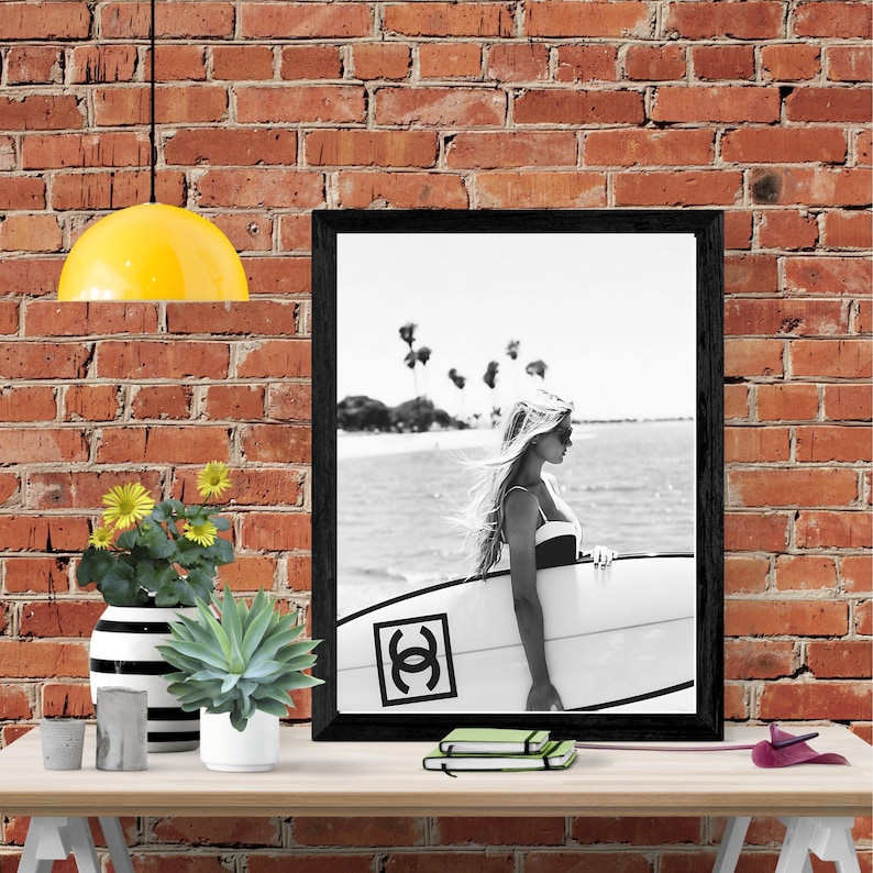 K&ouml;nnte beinhalten: Schwarzwei&szlig;foto einer Frau, die ein Surfbrett mit einem schwarzen "Chanel"-Logo an der Seite h&auml;lt. Die Frau tr&auml;gt einen Badeanzug und hat langes Haar. Das Foto ist in einem schwarzen Rahmen gerahmt und h&auml;ngt an einer Backsteinwand.