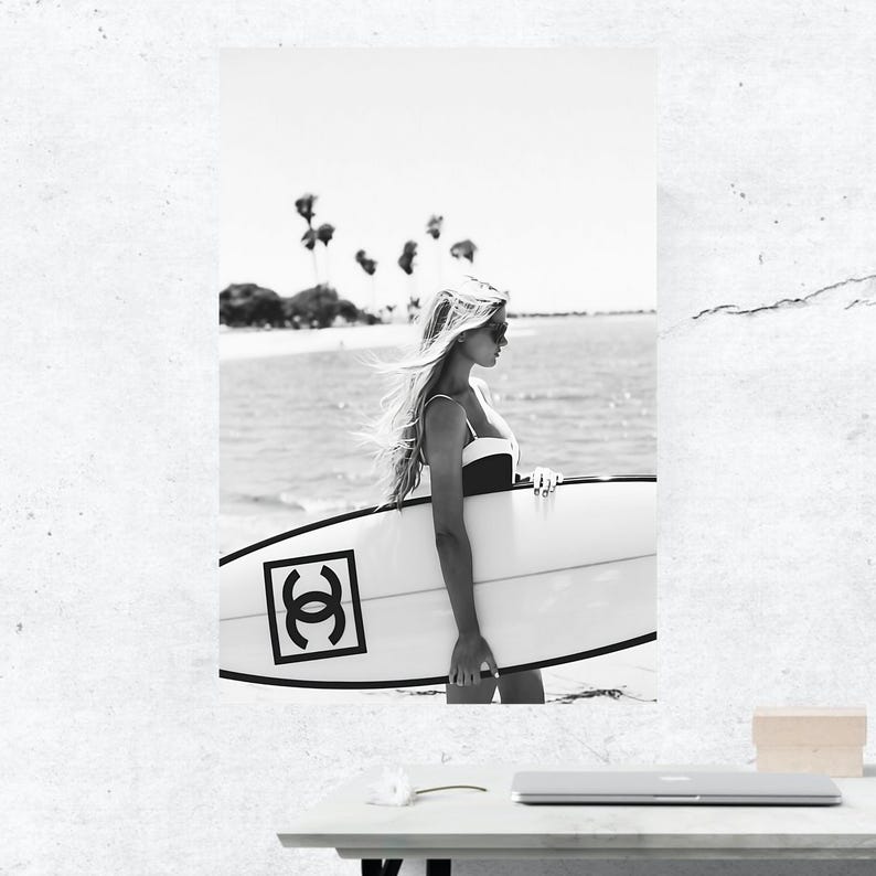 K&ouml;nnte beinhalten: Ein Schwarzwei&szlig;foto einer Frau im Badeanzug, die ein Surfbrett mit einem Chanel-Logo h&auml;lt. Die Frau steht an einem Strand mit dem Meer im Hintergrund.