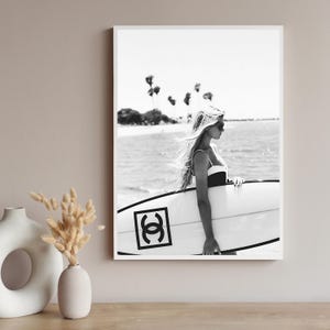 K&ouml;nnte beinhalten: Schwarzwei&szlig;foto einer Frau im Badeanzug, die ein Surfbrett mit Chanel-Logo h&auml;lt. Die Frau steht im Meer mit Palmen im Hintergrund.
