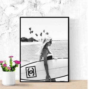K&ouml;nnte beinhalten: Schwarzwei&szlig;foto einer Frau im Badeanzug, die ein Surfbrett mit Chanel-Logo h&auml;lt. Die Frau steht an einem Strand mit Palmen im Hintergrund.
