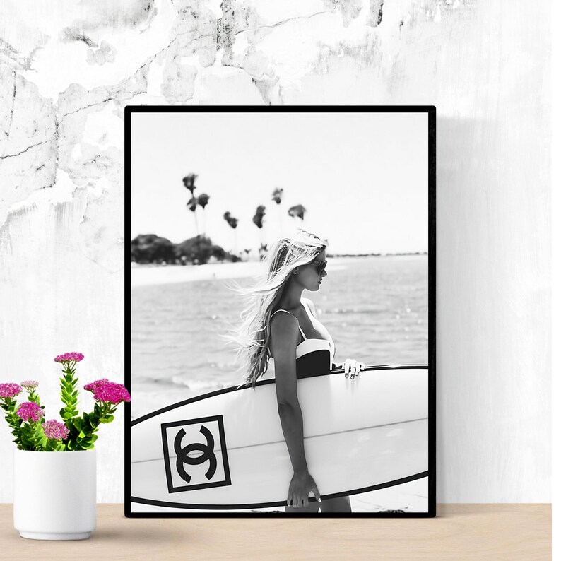K&ouml;nnte beinhalten: Schwarzwei&szlig;foto einer Frau im Badeanzug, die ein Surfbrett mit Chanel-Logo h&auml;lt. Die Frau steht an einem Strand mit Palmen im Hintergrund.