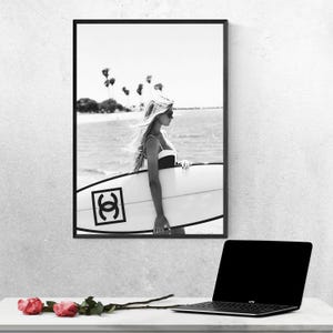 K&ouml;nnte beinhalten: Schwarzwei&szlig;foto einer Frau im Badeanzug, die ein Surfbrett mit Chanel-Logo h&auml;lt. Die Frau steht im Meer mit Palmen im Hintergrund.