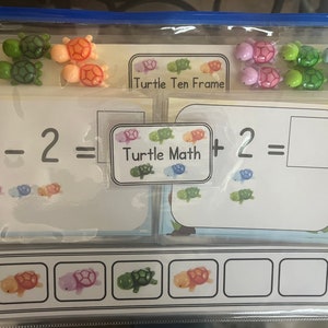 Può includere: Un sacchetto di plastica trasparente con una chiusura a zip contiene un set di manipolatori matematici a tema tartaruga colorati. I manipolatori includono un telaio di dieci, una carta di equazione matematica e un set di tartarughe individuali. La carta di equazione dice "3 - 2 = Turtle Math + 2 = ".