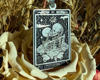 May include: Silver pendant necklace featuring a tarot card design. The rectangular pendant depicts two skeletons embracing, surrounded by roses, stars, and the text "THE LOVERS". The necklace is displayed against a backdrop of dried flowers and a rose.