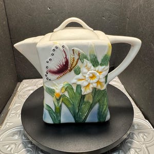 Puede incluir: Tetera de cerámica blanca con diseño de mariposa y flores. La tetera tiene un cuerpo cuadrado, un asa y una tapa. La mariposa tiene detalles en rojo, blanco y negro, y las flores son blancas y amarillas.
