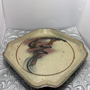 May include: A speckled ceramic plate with a unique, irregular shape. The plate features a cream-colored center with a brown rim and a decorative floral design in shades of brown, black, and pink. The plate has a slightly raised edge.