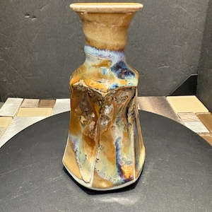 Può includere: Un vaso in ceramica con una smaltatura testurizzata e iridescente in tonalità di marrone, blu e oro. Il vaso ha una base larga e un collo stretto.
