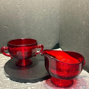 May include: A red glass creamer and sugar bowl set. The creamer has a handle and spout, while the sugar bowl has two handles. Both pieces have a ribbed design and sit on a black surface.