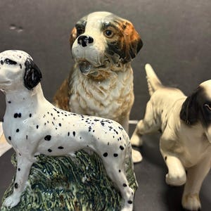 May include: Three ceramic dog figurines. A Dalmatian with black spots stands on a green base. A Saint Bernard with brown and white fur sits behind it. An English Setter with black and white markings is to the right.