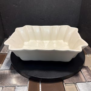 Peut inclure: Un plat à four en céramique blanche avec un bord festonné. Le plat est posé sur une surface noire.