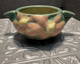 May include: A green and pink glazed ceramic bowl with a floral design. The bowl has two handles on the sides and is decorated with raised flowers.